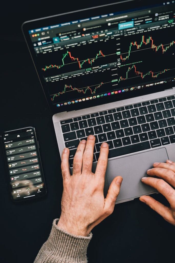 Hands Typing On A Laptop With Cryptocurrency Charts And Smartphone Display In View, Focusing On Digital Finance.